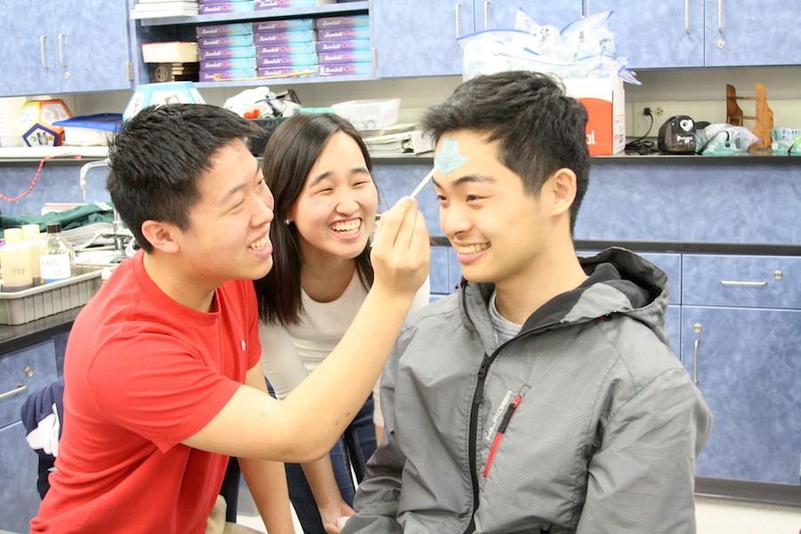 Junior Andrew Huang draws a blue arrow on senior Tim Chas head as senior Joo Kang looks on during chemistry teacher Robin Taylors Face Painting.