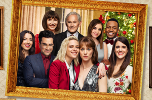 The cast of “Happiest Season” in front of a Christmas tree, with the stars Kristen Stewart and Mackenzie Davis in the very front. 