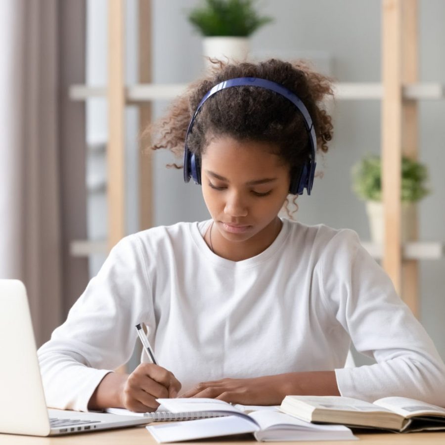 A girl takes notes while listening to music, effectively remaining focused on her current task. This use of audio stimuli for concentration is a typical depiction of the modern student.